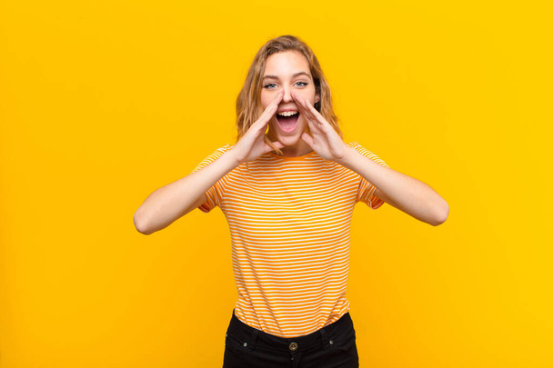 young blonde woman feeling happy, excited and positive, giving a big shout out with hands next to mouth, calling out against flat color wall - Foto, afbeelding