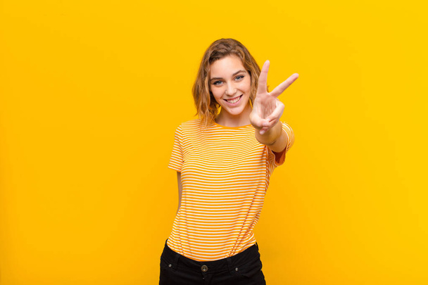 young blonde woman smiling and looking happy, carefree and positive, gesturing victory or peace with one hand against flat color wall - Fotoğraf, Görsel