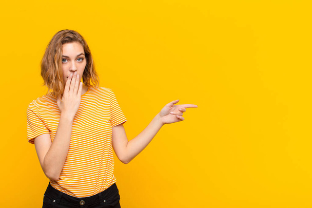 young blonde woman feeling happy, shocked and surprised, covering mouth with hand and pointing to lateral copy space against flat color wall - Foto, Bild