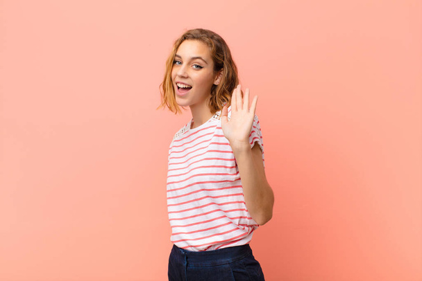 young blonde woman smiling happily and cheerfully, waving hand, welcoming and greeting you, or saying goodbye against flat color wall - Photo, Image