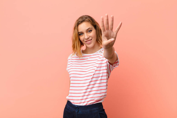 young blonde woman smiling and looking friendly, showing number four or fourth with hand forward, counting down against flat color wall - Photo, Image