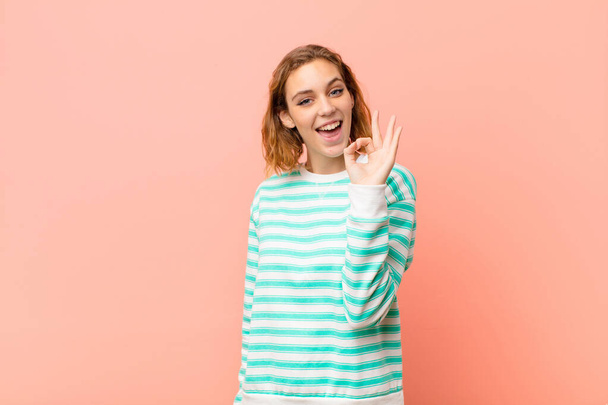 young blonde woman feeling successful and satisfied, smiling with mouth wide open, making okay sign with hand against flat color wall - Φωτογραφία, εικόνα