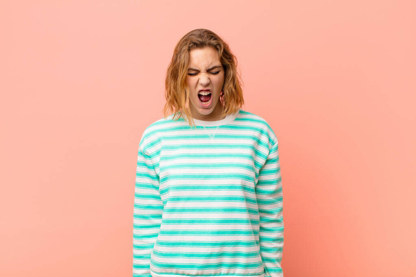young blonde woman shouting aggressively, looking very angry, frustrated, outraged or annoyed, screaming no against flat color wall - Fotoğraf, Görsel