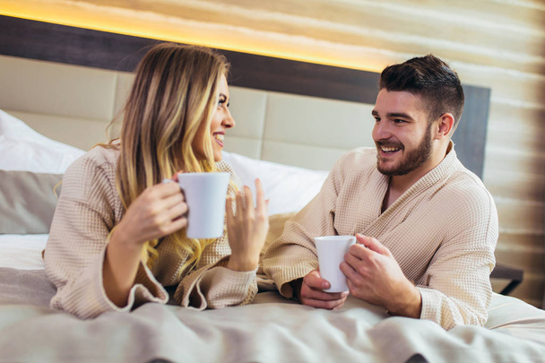 Jeune couple souriant vêtu de peignoirs couchés sur le lit et boire
 - Photo, image