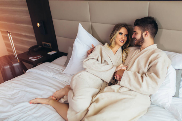 Ζευγάρι με μπουρνούζια ξαπλωμένο στο κρεβάτι στο δωμάτιο του ξενοδοχείου. - Φωτογραφία, εικόνα