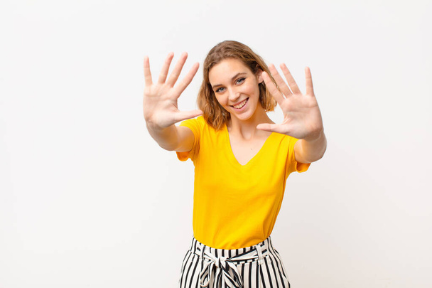 young blonde woman smiling and looking friendly, showing number ten or tenth with hand forward, counting down against flat color wall - Foto, Imagem