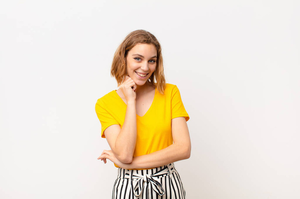young blonde woman smiling, enjoying life, feeling happy, friendly, satisfied and carefree with hand on chin against flat color wall - Foto, Imagem