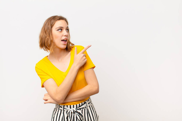 young blonde woman looking excited and surprised pointing to the side and upwards to copy space against flat color wall - Foto, Bild
