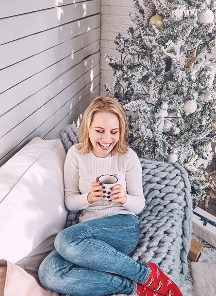 Beautiful young blonde woman is sitting and laughs on a sofa with a white mug in her hands on the background of a decorated New Year tree. Girl resting during the Christmas holidays - Foto, Imagem