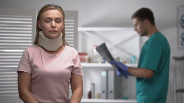 Sad female in foam cervical collar looking at cam, doctor examining x-ray result - Felvétel, videó