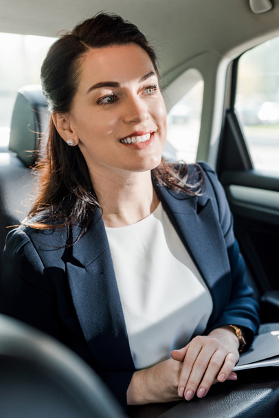 selective focus of happy woman in formal wear sitting in car with clenched hands  - 写真・画像