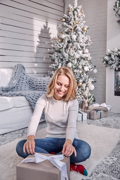 Gyönyörű szőke lány ül egy skandináv stílusú szobában díszített karácsonyra, és tart egy ajándék doboz egy íj a kezében. A lány ajándékkal nyit ki egy dobozt. Egy nő becsomagol egy ajándékot egy dobozba egy szalaggal.. - Fotó, kép