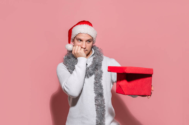 Man in een witte trui en rode Kerstman hoed op zoek naar de rode doos om erachter te komen wat geschenken zijn in. - Foto, afbeelding