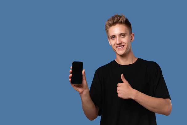 Blonder Teenager in schwarzem T-Shirt hält Smartphone mit Bildschirm mit erhobenem Daumen vor blauem Hintergrund in die Kamera. - Foto, Bild