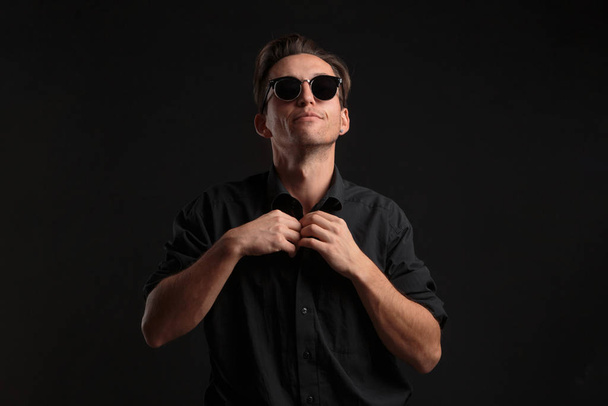 Confident stylish bussinesman in black eyeglasses buttoning up balck shirt isolated over black background. - Photo, Image