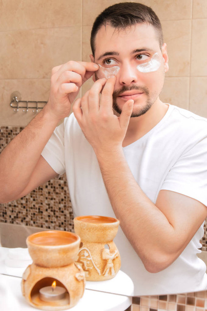 Um jovem olha-se ao espelho com manchas de prata debaixo dos olhos. Um tipo bonito na casa de banho faz-se uma máscara. Cuidados pessoais. Rotina matinal
. - Foto, Imagem