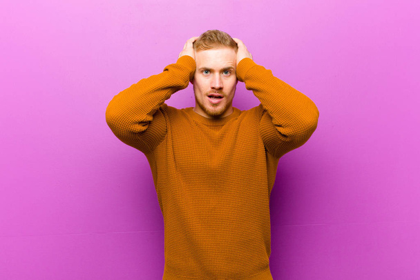 young blonde man wearing a jumper raising hands to head, open-mouthed, feeling extremely lucky, surprised, excited and happy - Foto, afbeelding