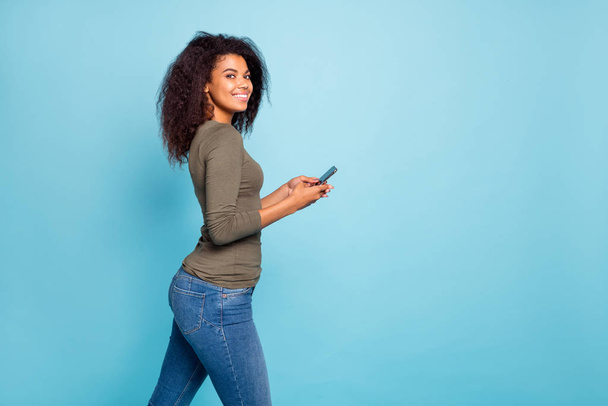Profile side photo of positive cheerful mulatto girl use her smartphone have fun on weekends enjoy read blog feednews in social network wear green sweater denim jeans isolated blue color background - Foto, immagini