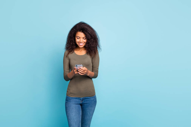 Portrait of positive cheerful focused blogger mulatto girl using her smartphone read feednews type feedback on blogs wear green sweater denim jeans isolated over blue color background - Fotografie, Obrázek