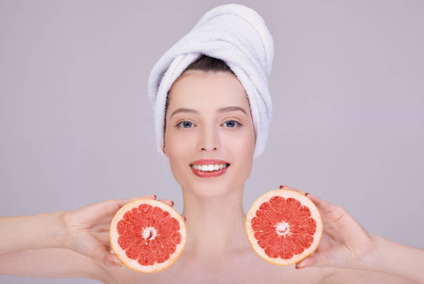 A smiling girl in a white towel on her head holds a cut grapefruit near her face. - Zdjęcie, obraz