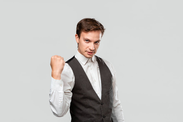 Zelfverzekerde zakenman in een wit overhemd en grijs vest met een gebaar van succes geïsoleerd over grijze achtergrond. - Foto, afbeelding