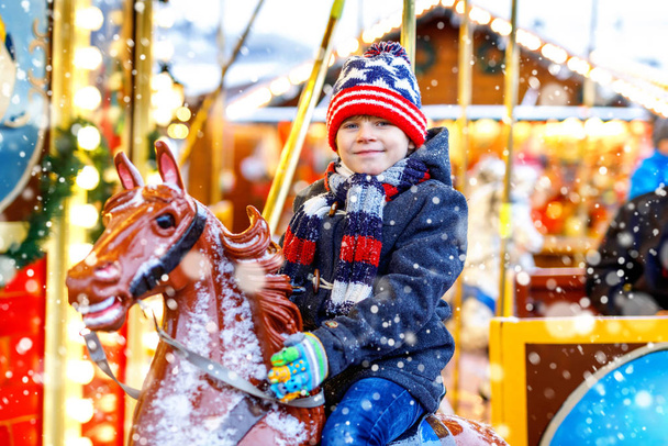Adorable little kid boy riding on a merry go round carousel horse at Christmas funfair or market, outdoors. Happy child having fun on traditional family xmas market in Cologne, Germany - Photo, Image