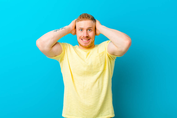 jonge blonde man draagt geel t-shirt gevoel van stress, bezorgd, angstig of bang, met handen op het hoofd, in paniek bij vergissing - Foto, afbeelding