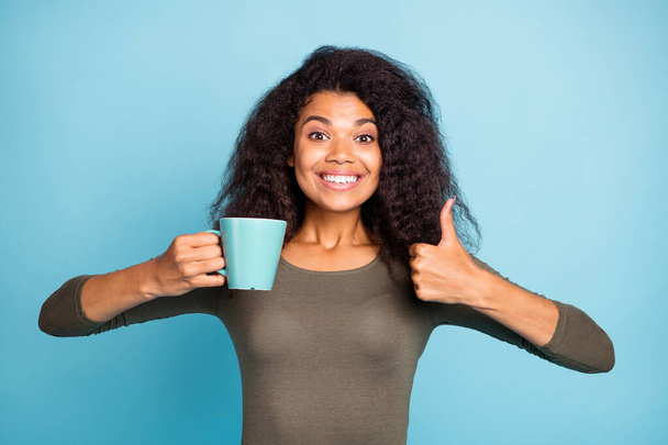 This cafeteria great. Portrait of positive cheerful dark skin girl hold cup hot caffeine beverage show thumb-up give feedback about excellent coffee wear green pullover isolated blue color background - 写真・画像