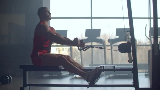Jongeman doet workouts op een rug met krachttrainingsmachine in een fitnessclub. Op atletische man doen workouts op een rug met power oefening machine in een fitnessruimte op de achtergrond van grote Windows - Video