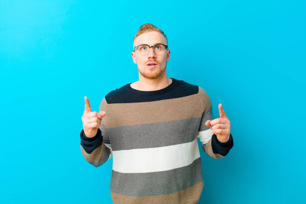 young blonde man feeling awed and open mouthed pointing upwards with a shocked and surprised look against blue background - Photo, image