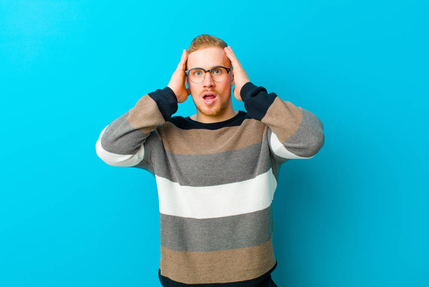 young blonde man looking unpleasantly shocked, scared or worried, mouth wide open and covering both ears with hands against blue background - Photo, image