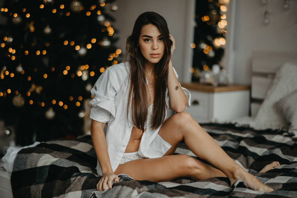 Giovane donna si siede sul letto sullo sfondo dell'albero di Natale
 - Foto, immagini