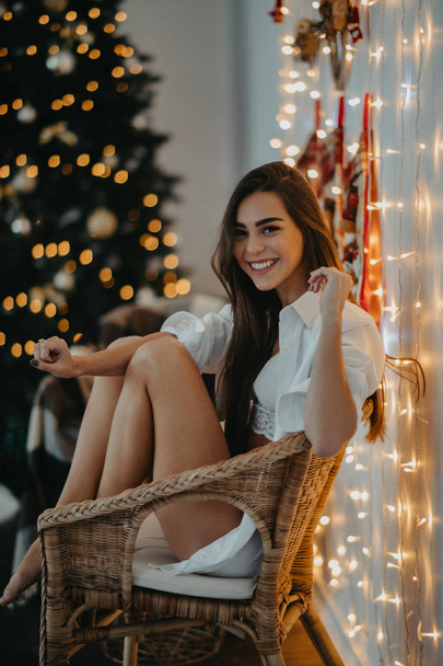 Jonge vrouw zit in de fauteuil en glimlacht tegen de kerstboom - Foto, afbeelding