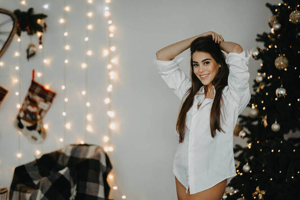 jonge vrouw staat en glimlacht tegen de achtergrond van kerstboom - Foto, afbeelding