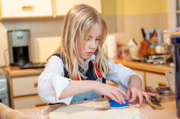 mooi jong blond meisje snijden uit deeg terwijl bakken in een rommelige keuken - Foto, afbeelding