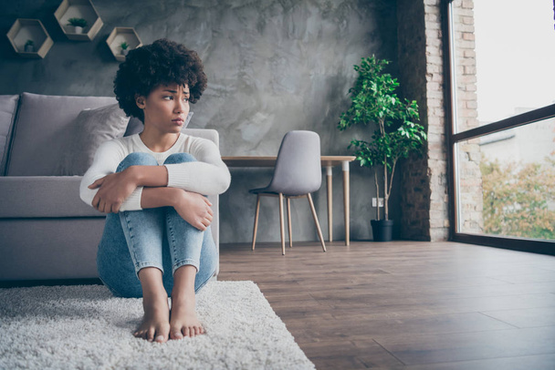 Photo of pretty dark skin curly lady sitting floor near sofa sad missing boyfriend looking window offended wear casual sweater jeans outfit flat indoors - Foto, Imagem