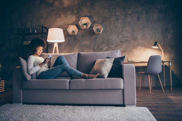 Profile photo of pretty dark skin curly lady domestic atmosphere texting telephone lying comfy couch in lamp light wear casual outfit evening living room indoors - Foto, immagini