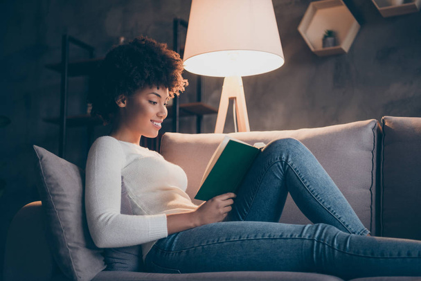 Photo of attractive dark skin curly lady holding favorite historic novel reading excited lying cozy couch wear casual sweater jeans outfit evening living room indoors - Zdjęcie, obraz