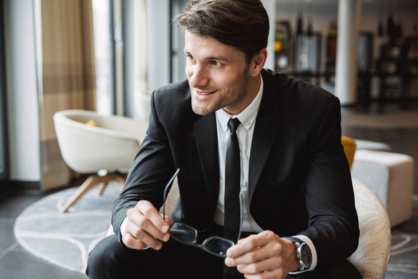 Photo of masculine businessman holding eyeglasses and sitiing on - Фото, изображение