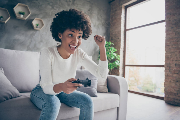 Photo of funny dark skin curly lady playing video games addicted want winning hold joystick triumphing football match sitting cozy couch wear casual sweater jeans outfit flat indoors - 写真・画像