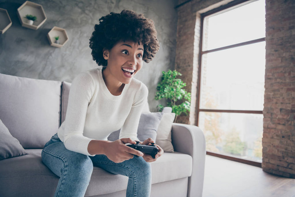 Photo of funny dark skin curly lady playing video games addicted want winning hold joystick sitting comfortable couch wear casual sweater jeans outfit flat indoors - Valokuva, kuva