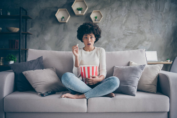 Photo of pretty dark skin wavy lady eating popcorn watching frightening horror film open mouth scary moment sitting cozy couch casual sweater jeans outfit flat indoors - Foto, Imagem