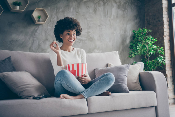 Photo of pretty funny dark skin wavy lady homey mood eating popcorn watching favorite humor television show sitting cozy couch casual sweater jeans outfit flat indoors - Foto, imagen
