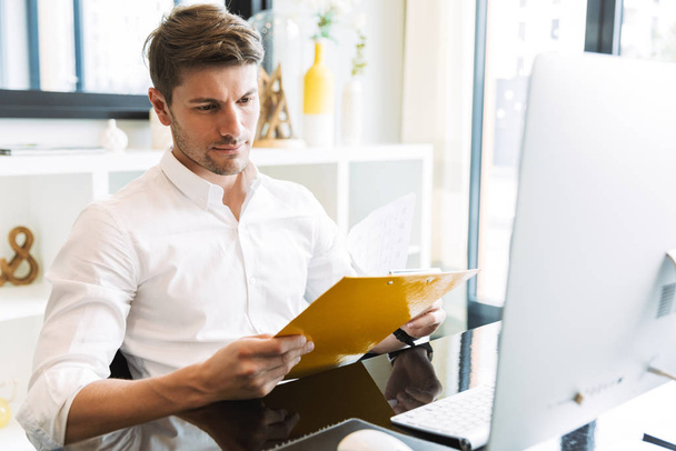 Image of handsome businesslike man holding clipboard while worki - Foto, afbeelding