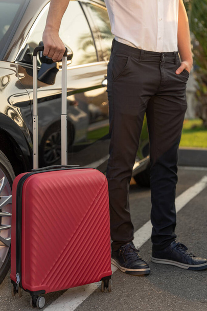 Un hombre con una maleta roja al lado del coche negro. El concepto de traslado al aeropuerto o taxi
 - Foto, Imagen