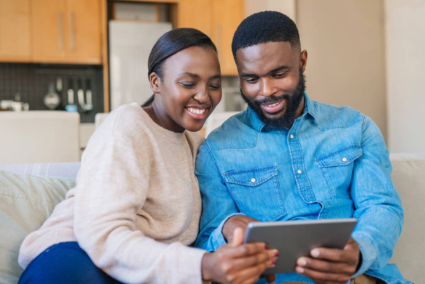 Smiling young African American couple using a digital tablet while relaxing together on their living room sofa at home - Fotó, kép