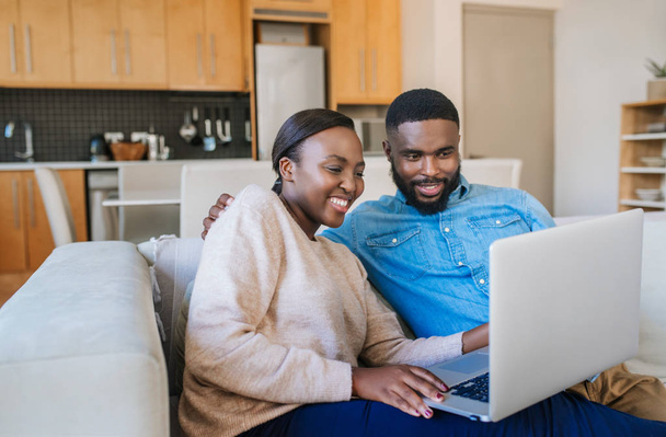 Laughing young African American couple browsing online with a laptop while sitting together in their living room at home - Foto, imagen