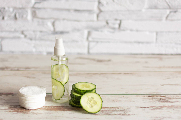 Zelfgemaakte verfrissende gezichtslotion in flesje met plakjes groene komkommer en wattenschijfjes op witte houten ondergrond. - Foto, afbeelding