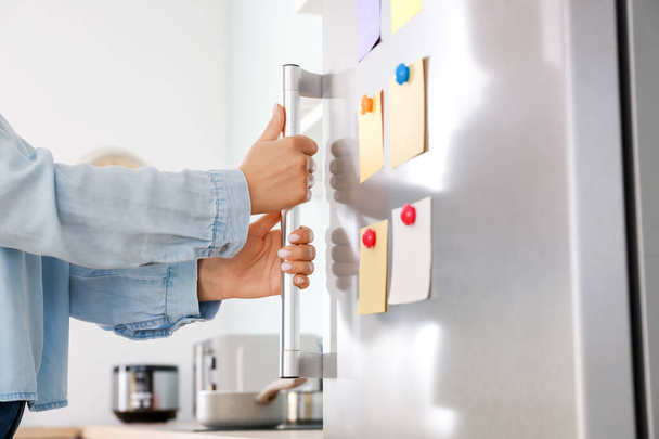 Woman opening modern fridge with stickers on door in kitchen - Foto, afbeelding