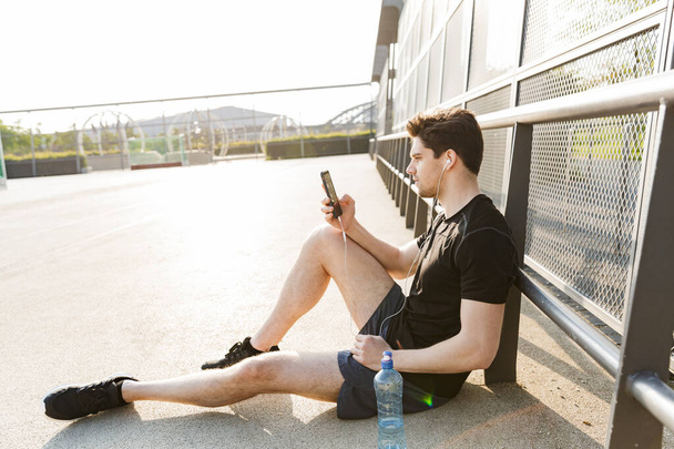Portrait of young man wearing earphones holding cellphone while  - Fotó, kép